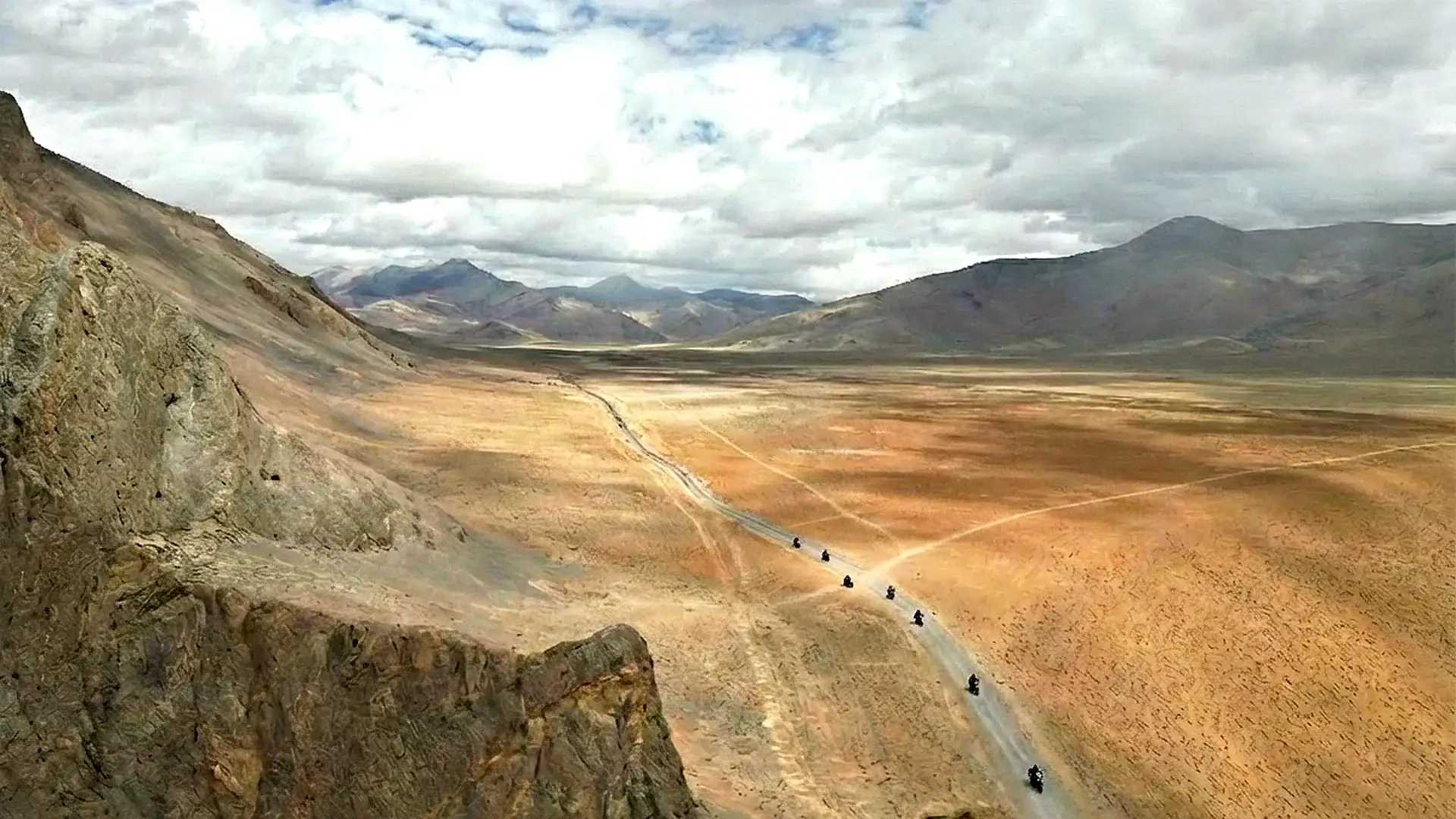 Long winding dirt road through open plains and mountains under cloudy sky.