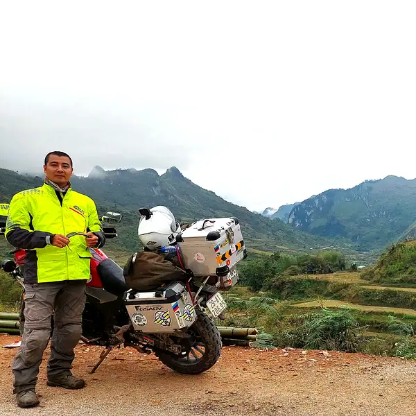 Motorcycle rider standing next to a touring bike with scenic mountain landscape in the background.